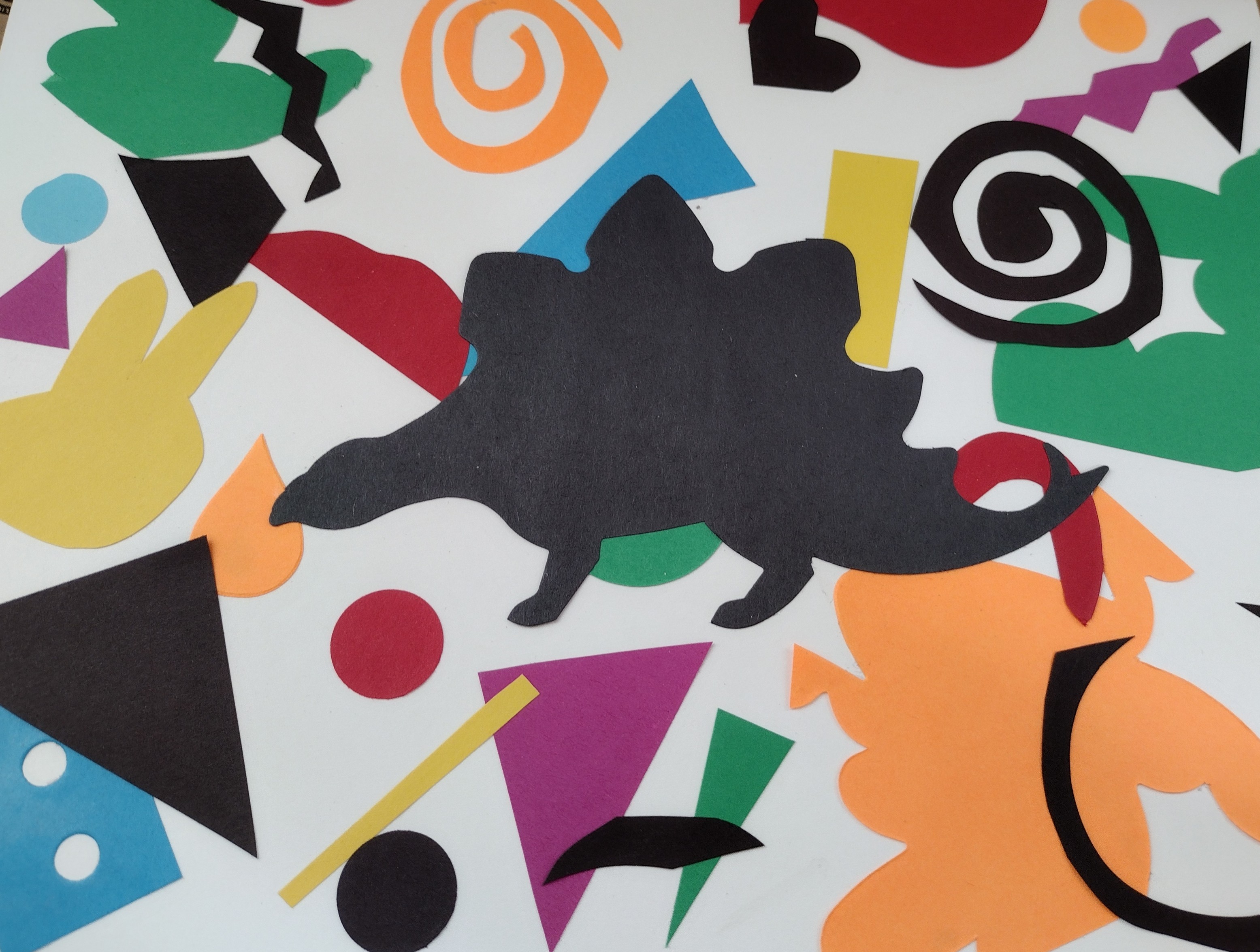 Various colorful shapes on a white piece of cardstock with a black dinosaur die cut in the foreground. 
