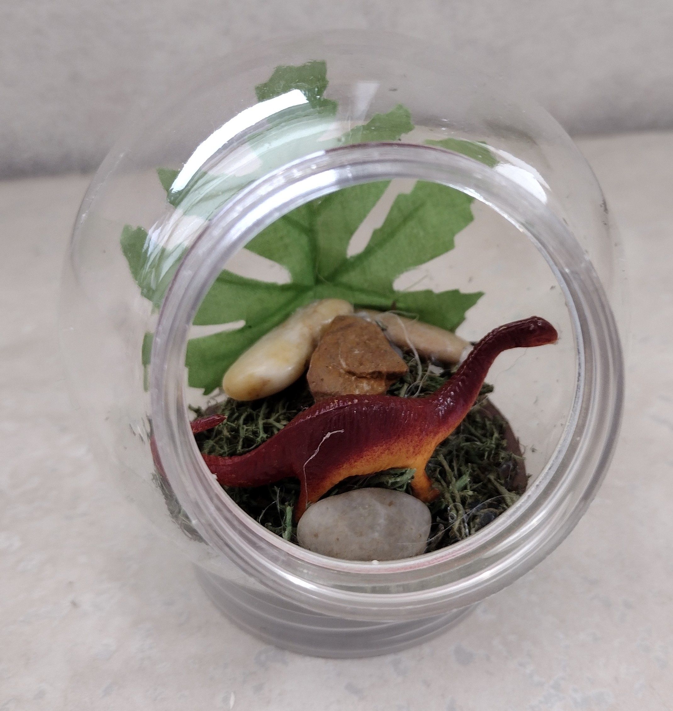 Plastic bulb with a toy dinosaur, rocks, and moss inside of it. 